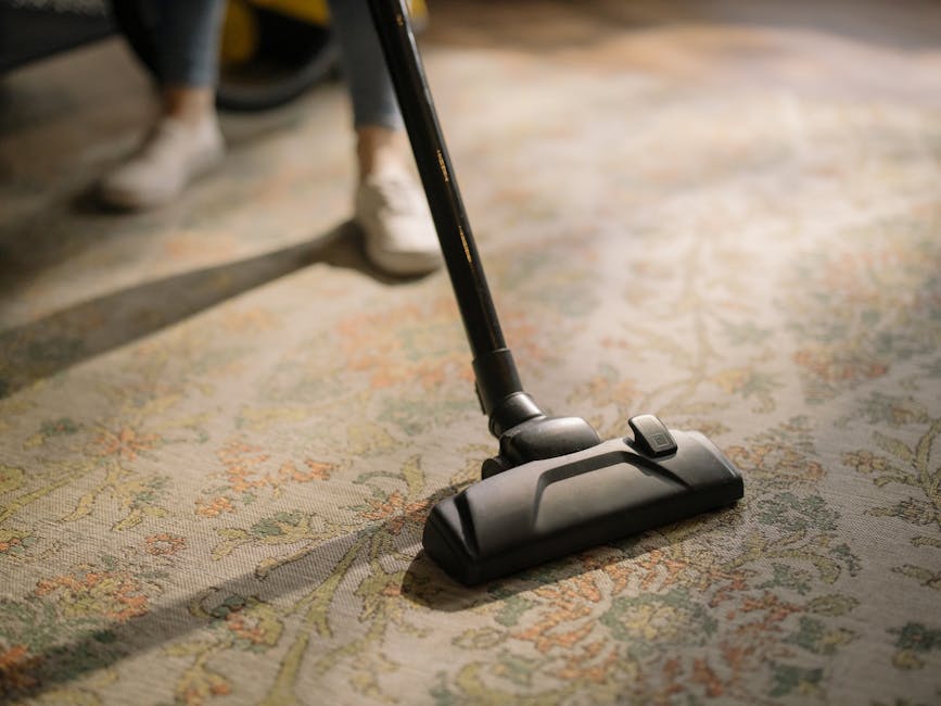 Close-up of a wet vacuum cleaner with a transparent dust container and a flexible, patterned hose attached to a floor cleaning head, which is positioned on a beige carpeted surface. The vacuum cleaner