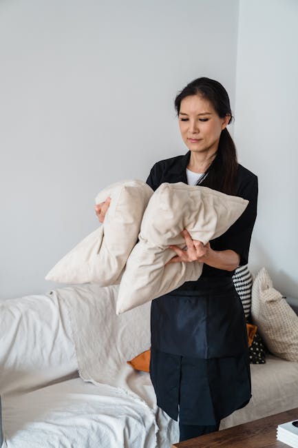 Close-up image of a person using a handheld vacuum cleaner to deep clean the fabric upholstery of a green lounge chair, with another dark gray fabric sofa visible in the background. The green chair fe